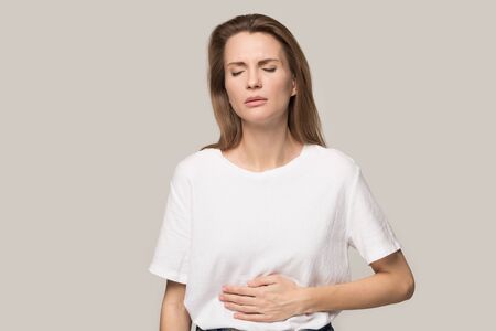 Unhealthy millennial girl isolated on grey studio background touch stomach suffer from indigestion or diarrhea, unhappy young woman feel sick ill touch belly have pain from gastritis or pancreatitisの写真素材