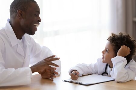 Cute little biracial boy in medical uniform play act as doctor talk with smiling african American pediatrician, funny small child patient have fun involved in funny game with family gp in hospitalの写真素材