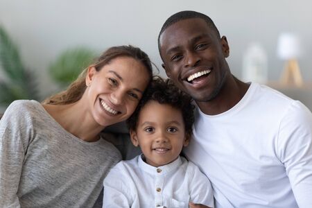 Portrait of happy young multinational family with preschooler biracial boy child posing at home, smiling international parents and mixed race ethnicity little son look at camera enjoy time together