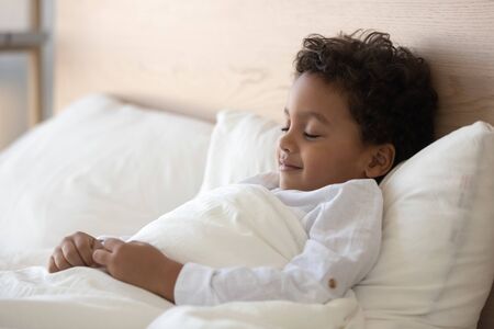 Peaceful cute little biracial boy sleeping in white cozy bed at home, happy smiling mixed race ethnicity preschooler child fall asleep relax in comfortable bedroom, having day rest daydreaming at homeの写真素材