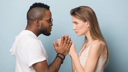 Multiracial couple isolated on blue studio background hold hands have intimate close moment together, loving multiethnic millennial man and woman look in eyes share secret, side profile viewの写真素材
