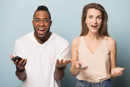 Excited multiethnic couple isolated on blue studio background look at camera shocked by amazing good...