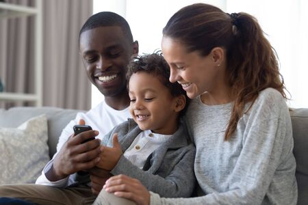 Happy international young family with little son sit on couch watching cartoons on cellphone together, smiling multiracial parents relax on couch at home with biracial boy child using smartphoneの写真素材