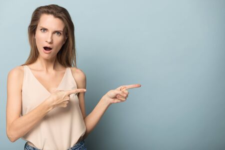 Surprised Caucasian millennial woman isolated on blue studio background point at blank copy space advertising spot, shocked young female show recommend good service or promotion dealの写真素材