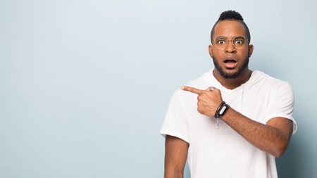 Shocked african American millennial man in glasses isolated on blue studio background point with finger at blank copy space aside, surprised biracial male show at amazing sale deal offer or promotionの写真素材