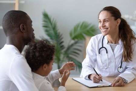 Smiling female doctor consult african American dad with little boy patient write prescription in medical card, happy positive woman pediatrician talk with biracial father with son visiting hospitalの写真素材