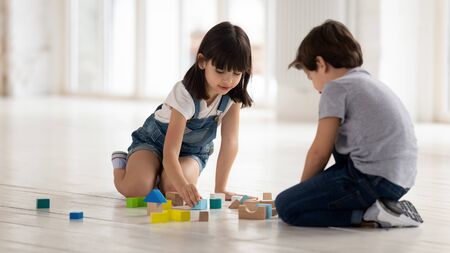 Happy little brother and sister sitting on heated warm floor, playing with wooden multicolored bricks, constructing building houses. Carefree siblings enjoying spending leisure time together at home.の写真素材