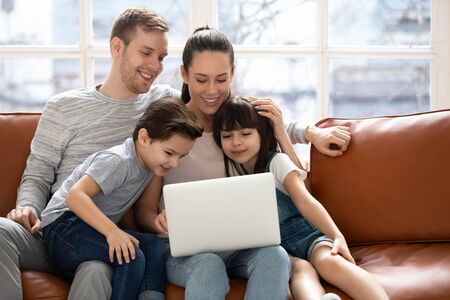 Joyful family of four sitting on couch, bonding embracing, looking at laptop screen, watching comedy movie or vacation photos, making purchases in popular online store, shopping together at home.の写真素材