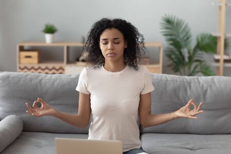 Peaceful african American millennial girl distracted from computer work sit on couch at home meditating, calm focused black young woman practice yoga on sofa in living room. Stress free conceptの写真素材
