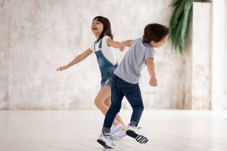 Full length happy little boy and girl holding hands, twisting, running, playing together, laughing. Energetic excited siblings having fun at home, engaged in indoors activity, feeling overjoyed.の写真素材