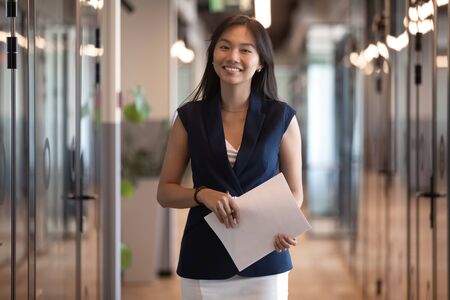 Portrait of smiling Asian businesswoman holding papers, document, attractive woman standing in hallway, smiling female employee looking at camera, confident executive posing in modern officeの写真素材