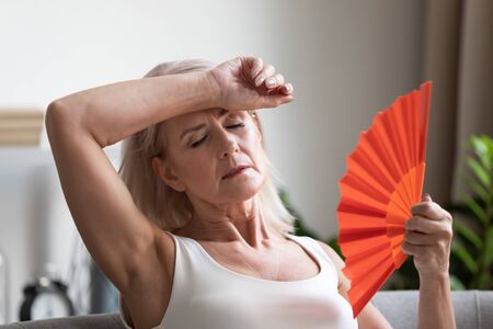 Tired overheated middle aged lady wave fan suffer from menopause exhaustion complain on heat at home, stressed old woman sweat feel uncomfortable hot in summer weather problem without air conditionerの写真素材