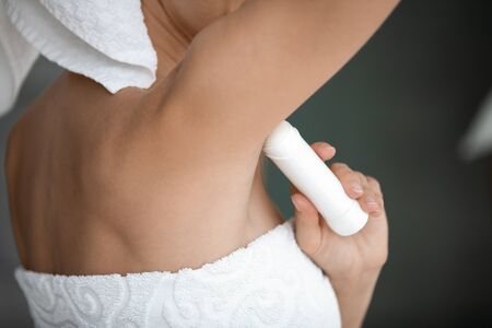 Young lady wrapped in white towel raise arm apply antiperspirant in armpit, woman use underarm deodorant stick for daily hygiene everyday freshness, hyperhidrosis treatment concept close up rear viewの写真素材