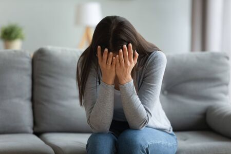 Sad depressed young adult woman crying alone at home, upset desperate girl in tears trouble hiding face in hands feeling stressed ashamed worried about problems regret abortion concept sit on sofaの写真素材