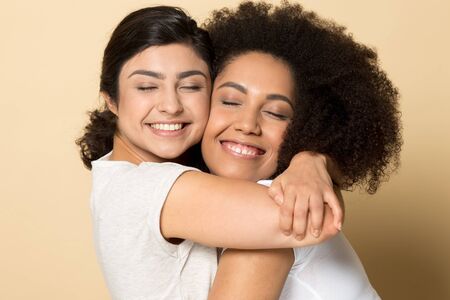 Smiling diverse girls with closed eyes, best friends hugging close up, standing isolated on brown studio background, two young female expressing love and support, true friendship, good relationsの写真素材