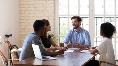 Happy young male manager executive in eyewear having fun, laughing during brainstorming meeting with diverse colleagues. Smiling mixed race group of coworkers listening to boss joking at office.の写真素材