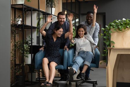 Overjoyed young diverse colleagues having fun, riding forward on chairs on coworking office. Happy international team laughing, enjoying together teambuilding funny activity at modern workplace.の写真素材