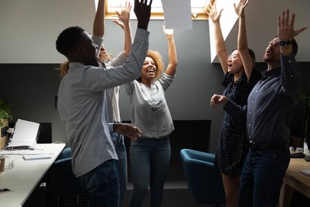 Euphoric young mixed race startup business team throwing papers in air, celebrating successful project finish. Overjoyed millennial diverse coworkers feeling proud for excellent team work results.の写真素材