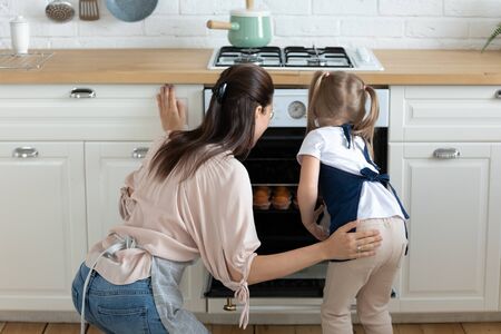 Back rear view young mommy and curious little kid girl wearing aprons, looking into oven, checking if muffins ready to eat. Happy family enjoying pastry cooking process together at modern kitchen.の写真素材