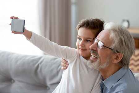 Happy grandfather wearing glasses and little grandchild taking selfie on phone, smiling granddad and...