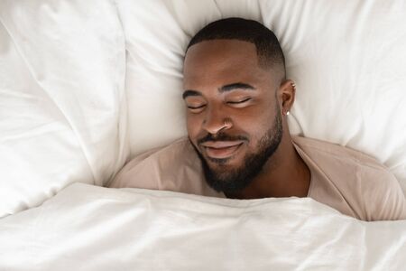 Top view of happy african American man sleeping in comfortable white bed seeing good pleasant dreams, calm biracial male feel fatigue resting napping in cozy bedroom under linen bedding sheetsの写真素材