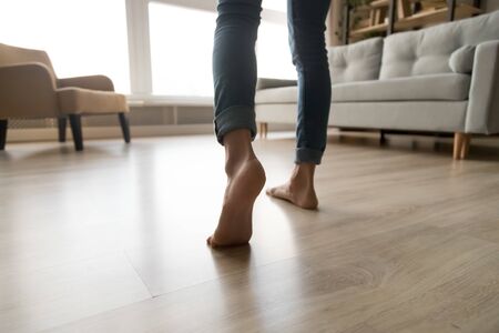 Close up of barefoot young woman in jeans feel comfortable walking on warm heated wooden floor at...