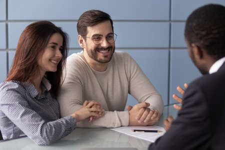 Happy Caucasian millennial couple listen to african American realtor or broker talking explain...