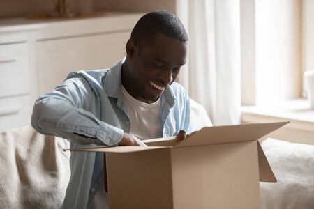 Happy african American man sit on couch in living room unpack box excited with successful online...