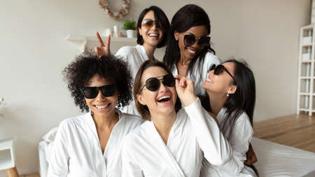 Cheerful young ladies laughing look at camera wear sunglasses bathrobes, happy multi ethnic women best friends having fun celebrate bridal shower bachelorette hen party at home hotel bedroom togetherの写真素材