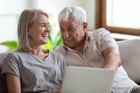 Overjoyed mature woman sitting on sofa with joyful elder man, holding computer on knees. Happy married family spouse laughing, having fun, joking, watching funny movies or videos on laptop at home.の写真素材