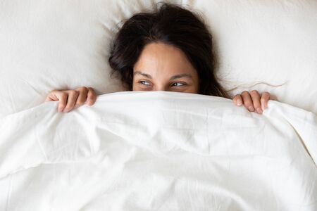 Above close up view young millennial cheerful woman lying under blanket inside of cozy bed on white...