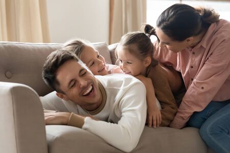 Cheerful happy parents laughing playing with small adorable kids daughters lying on couch, smiling mom dad and cute little preschool children girls having fun enjoy family lifestyle on cozy weekendの写真素材