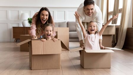 Excited young family having fun on moving day, happy parents pushing cardboard boxes with funny cute...