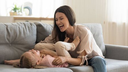 Cheerful family young adult mother tickling cute small child daughter laughing playing together on sofa in house living room, happy mom and little preschool kid having fun cuddle enjoy lifestyle gameの写真素材