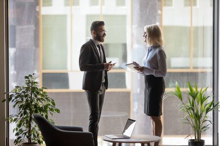 Young Arabic and middle-aged Caucasian businesspeople stand in modern office talk discussing ideas, millennial Arabian man employee speak consider cooperation with woman business partnerの写真素材