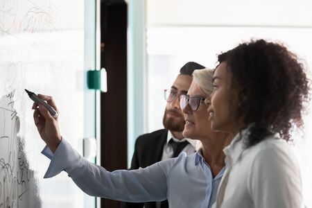 Side view of diverse multiethnic colleagues write draw on white board in office discussing company startup project or plan, multiracial employees brainstorm engaged in team creative thinkingの写真素材