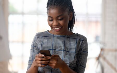 Close up head shot portrait smiling young african american business lady holding smartphone, using corporate software applications, texting email to clients, chatting in messenger with partners.の写真素材