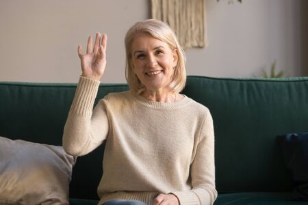 Attractive elderly woman sitting on couch at home looking at camera makes video call waving hand...