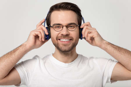 Smiling Caucasian millennial man in glasses wear blue modern headphones look at camera excited with device, happy guy in spectacles listen to music enjoy quality sound in new earphonesの写真素材