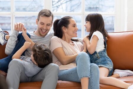 Playful young parents have fun tickle cute small kids daughter and son enjoy weekend at home, happy...