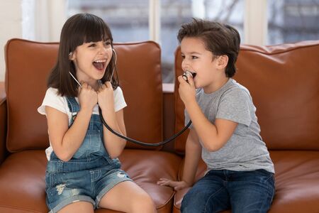 Cute little boy and girl sit on couch laugh play doctor and patient game using stethoscope, smiling small preschooler siblings feel overjoyed engaged in funny activity, have fun together at homeの写真素材