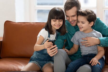 Happy young father and cute little kids sit on couch watch cartoons on smartphone together, smiling dad parent spend time relax with small preschooler son and daughter have fun using cellphone playingの写真素材