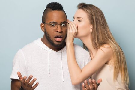 Woman whispering in African American man ear, telling unexpected news, shocked surprised boyfriend hearing unbelievable rumors from girlfriend, friends gossiping, isolated on studio backgroundの写真素材