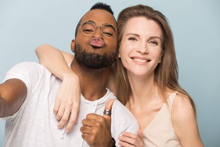 Funny African American man and Caucasian woman taking selfie together, looking at camera, showing thumbs up and kiss, smiling diverse couple embracing, standing isolated on studio backgroundの写真素材