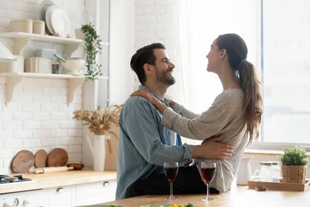 Happy young couple drinking red wine talking bonding in modern kitchen, smiling affectionate husband and wife laughing celebrating romantic anniversary together embracing enjoy married life at homeの写真素材