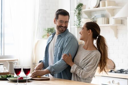 Happy positive married young couple husband and wife cutting vegetable salad embracing talking laughing having fun cooking together preparing healthy meal enjoy cozy romantic lifestyle with red wineの写真素材