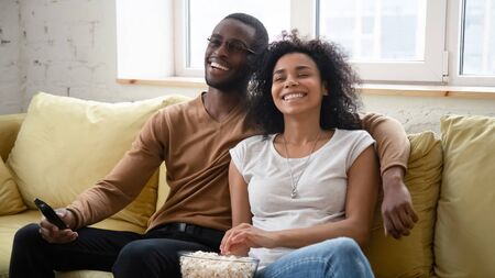 Overjoyed african American couple sit relax on cozy sofa in living room watch TV eating popcorn,...