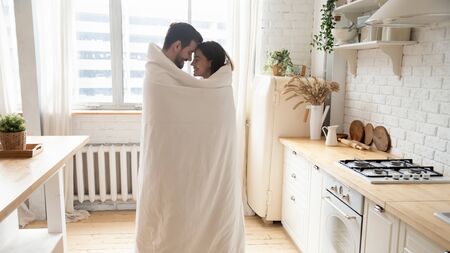 Young affectionate couple standing in modern kitchen wrapped with warm blanket, happy romantic husband and wife bonding enjoying cozy morning at home feeling love having fun covered with plaidの写真素材