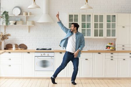 Carefree funny young man having fun dancing alone in modern kitchen interior, active happy funky single guy enjoying silly movements dance standing at home listening music celebrating freedom conceptの写真素材