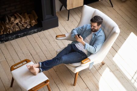 Relaxed casual guy lounge sit on comfortable armchair in modern house room with wooden floor fireplace using smartphone apps, happy young man hold cell phone play mobile games at cozy home, top viewの写真素材